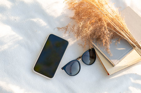 Still life with smartphone, books, dried flowers, and sunglasses on a white blanket in warm natural light, evoking a peaceful weekend vibeの写真素材