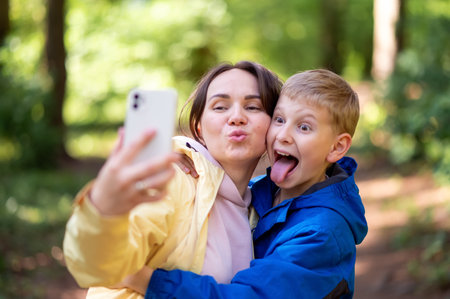 Happy mother and son taking a selfie in a spring park, boy making faces and sticking out his tongueの写真素材