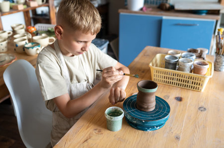 Blond boy carefully painting handmade clay cup, creative kids activity in cozy home pottery workshopの写真素材