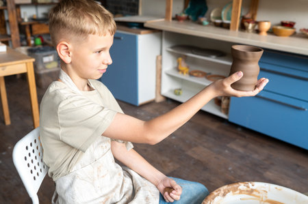 Boy sitting in pottery studio, smiling while holding freshly crafted clay cup, child's creative hobbyの写真素材