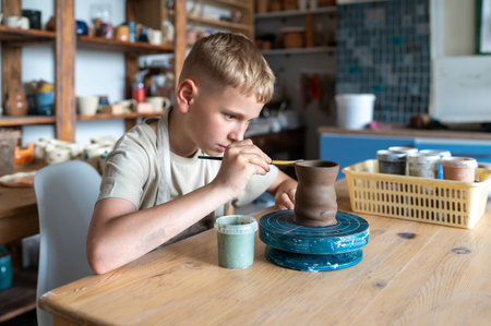 Blond boy carefully painting handmade clay cup, creative kids activity in cozy home pottery workshopの写真素材