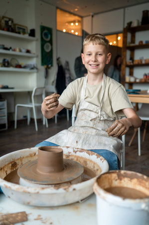 Blond boy learning pottery, close-up of hands shaping clay on a wheel, creative kids activity in cozy pottery studio, teamwork and manual craft skills conceptの写真素材