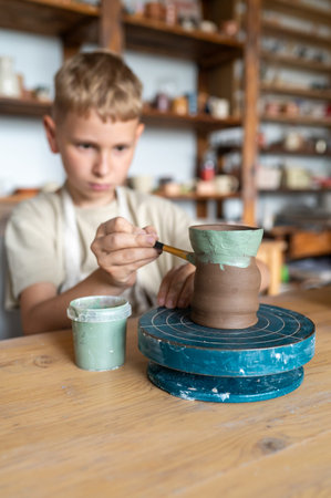 Blond boy carefully painting handmade clay cup, creative kids activity in cozy home pottery workshop