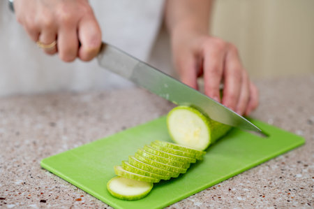 Close-up of hands slicing zucchini with a knife on a green board, concept: fresh meal preparation at...