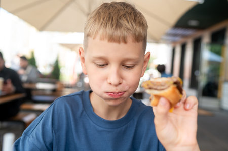 Teenage blond boy enjoying time at outdoor cafe with a burger, peaceful summer day, casual and friendly atmosphere in natural lightの写真素材