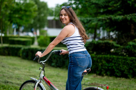 Joyful middle-aged woman cycling along a green park path on a warm summer day, enjoying fresh air, nature, and active outdoor lifestyle with a bright smileの写真素材