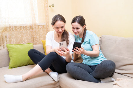 Two female friends laughing on the couch while looking at a smartphone, discussing something funny,...