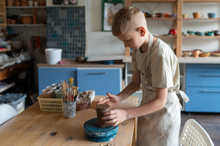 Happy boy shaping wet clay at pottery wheel in cozy studio, learning traditional craft skills