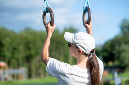 Young woman in cap and sunglasses training on gymnastic rings at outdoor sports ground, concept of...