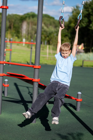 Blond teenage boy swinging on gymnastic rings at outdoor playground, concept of fun, strength,...
