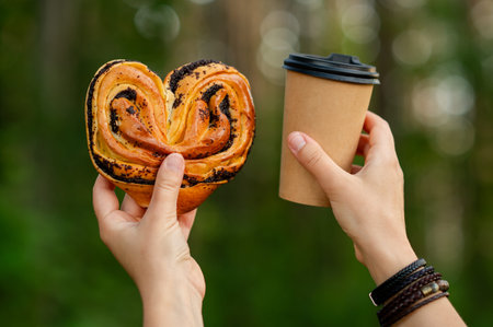 Woman's hands hold a paper cup with hot beverage and a sweet bun while enjoying a cozy picnic in the...