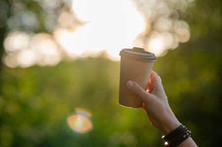 Woman's hand holds a paper cup with a hot beverage, surrounded by forest bathed in golden sunset...