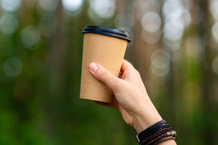 Woman's hand holds a paper cup with a hot beverage, surrounded by forest bathed in golden sunset light, evoking warmth and cozy autumn atmosphereの写真素材