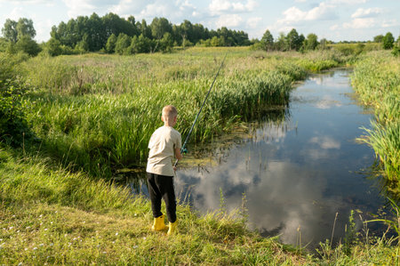 Child standing with fishing rod by calm river on summer day, enjoying outdoor family tripの写真素材