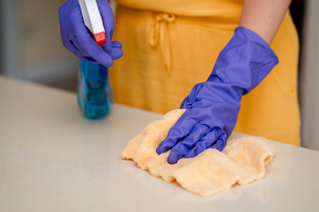 Woman wearing gloves sprays furniture polish and carefully wipes a wooden table with a cloth,...