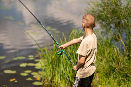 Happy blond boy standing on lake shore with fishing rod, fishing on bright summer day, enjoying...