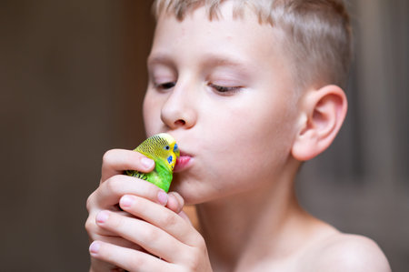 Blond teenage boy gently holding a green budgerigar and kissing it, concept of love, care and...