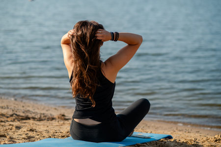 Back view of woman in sportswear sitting on the shore of a lake, looking into the distance, enjoying...