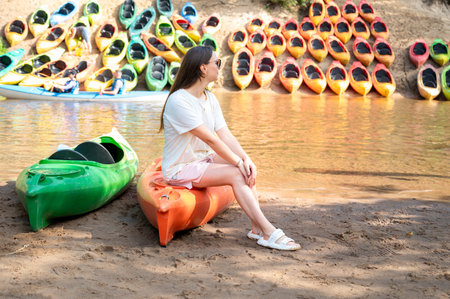 Woman wearing sunglasses sits on a kayak at the river shore, with numerous kayaks on the opposite bank, ready for summer rafting adventure, outdoors, water sport, natureの写真素材