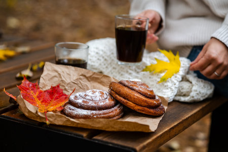 Only hands and torso of woman in white knitted sweater visible, holding cup of hot coffee next to pastries and bright maple leaves on wooden table in autumn parkの写真素材