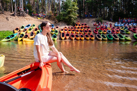 Young woman in casual outfit sits on a kayak on the river shore, surrounded by multiple kayaks, ready for a summer rafting adventure, outdoors, water sport, natureの写真素材