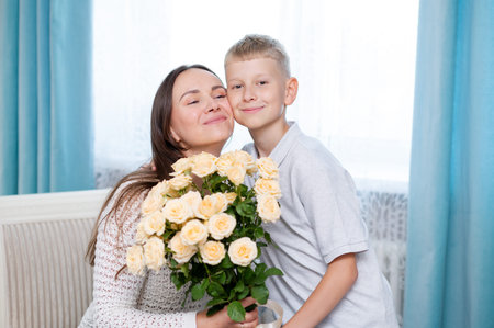 Teenage boy warmly hugs his mother while presenting a bouquet of yellow roses in a cozy room, celebrating Mother's Day with love and gratitudeの写真素材