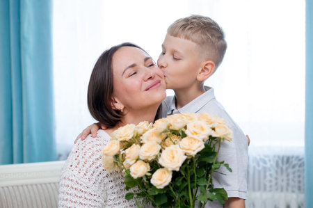 Teenage boy warmly hugs his mother while presenting a bouquet of yellow roses in a cozy room, celebrating Mother's Day with love and gratitudeの写真素材