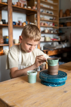Blond boy carefully painting handmade clay cup, creative kids activity in cozy home pottery workshopの写真素材