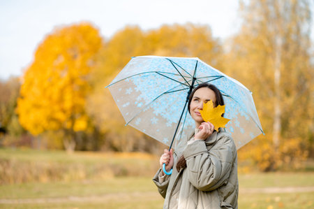 Conceptual autumn scene with smiling woman in park, holding maple leaf over eye beneath transparent umbrella, celebrating fall colors and playful moodの写真素材