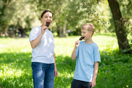 Happy mother and son enjoying chocolate ice cream on a stick together in sunny summer park, concept of family leisure and sweet treats outdoorsの写真素材