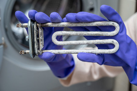 Person wearing purple cleaning gloves holds a used washing machine heating element covered with limescale near the open door, demonstrating part replacement or maintenance processの写真素材