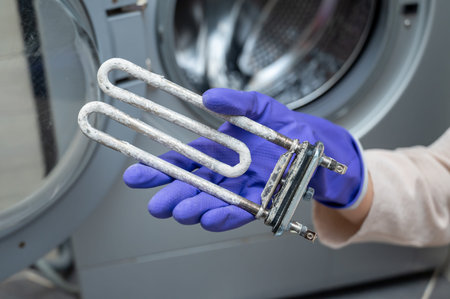 Person wearing purple cleaning gloves holds a used washing machine heating element covered with limescale near the open door, demonstrating part replacement or maintenance processの写真素材
