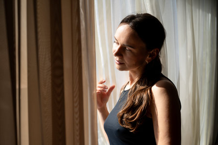 A young woman sits indoors, gazing thoughtfully out the window as warm sunlight falls on her,...