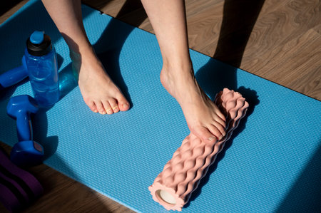 Woman legs performing self myofascial release massage on foam roller at home, with dumbbells and water bottle on yoga mat, concept of fitness recovery and healthy lifestyleの写真素材
