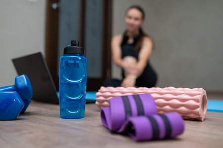 Dumbbells, resistance band and foam roller on yoga mat in the foreground, with athletic woman...