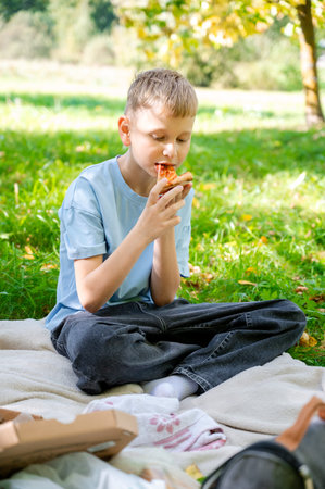 Blond teenage boy enjoying slice of pizza at summer picnic, outdoor leisure activity with tasty fast food, casual lifestyle scene in green park nature environmentの写真素材