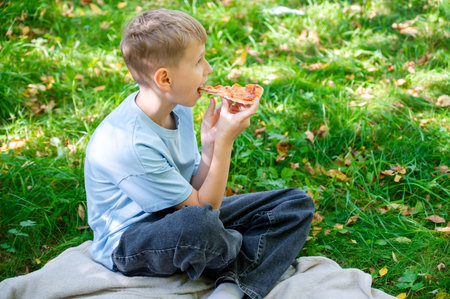 Blond teenage boy enjoying slice of pizza at summer picnic, outdoor leisure activity with tasty fast food, casual lifestyle scene in green park nature environmentの写真素材