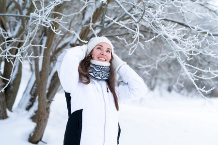 Smiling young woman adjusting knitted hat in snowy winter park. Cozy winter lifestyle, cold weather,...