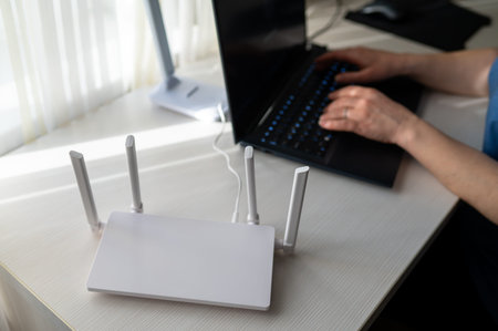 Top view of a modern internet router with four antennas on a white wooden table. Female hands are...