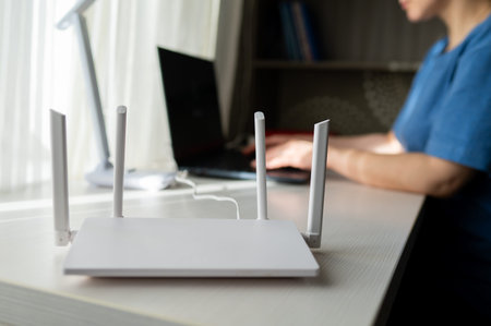 Close-up of a modern wireless router with antennas on a wooden desk. In the blurred background, a...