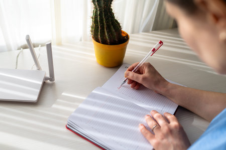 Close-up of woman hand writing in a red notebook on a bright desk with a white wireless router....