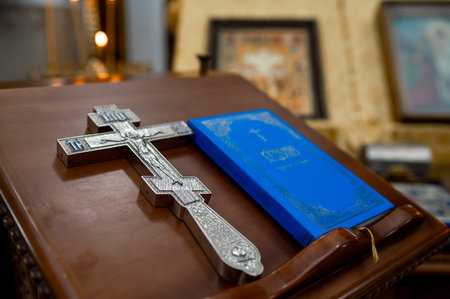 A high-quality close-up of a silver crucifix and a blue holy bible lying on a wooden stand in an orthodox church interior. The background features blurred icons and a warm candle light in a sanctuaryの写真素材