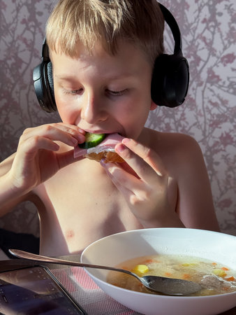 A young boy is having a meal at home wearing black wireless headphones. He is biting a sandwich with...