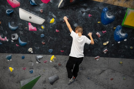 A schoolboy in a white t-shirt and black pants is training on a dark climbing wall. He is reaching for holds in a professional bouldering center during his sport lesson or active leisure at homeの写真素材