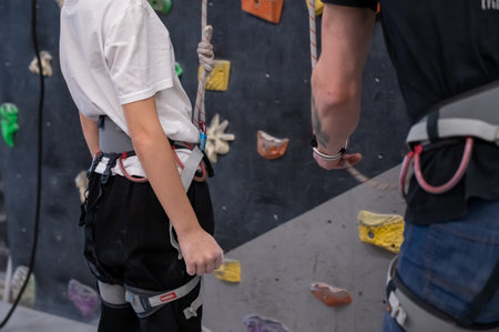 Close up of a child and a coach preparing for indoor rock climbing. The instructor is holding a rope...