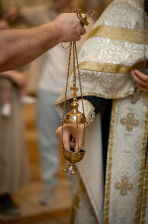 Close up of priest in white vestment with gold embroidery using brass censer for burning incense in...