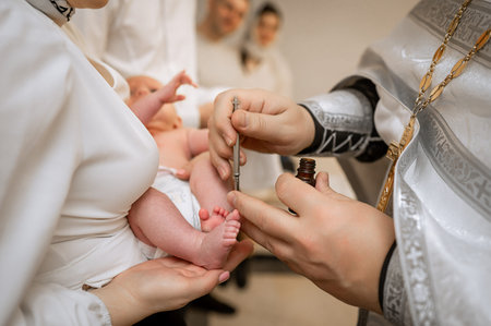 Close up of priest hands using metal brush for chrismation on infant leg held by mother during christening ritual in church religious tradition of anointing baby with oil and faithの写真素材