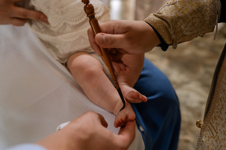 Close up of priest hands using wooden brush for chrismation on infant leg during christening ritual...
