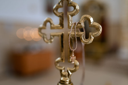 A close up shot of a tiny gold crucifix for a baby baptism ceremony hanging on a larger church vessel. The background features a soft blurred interior of a Christian temple with warm light and bokehの写真素材