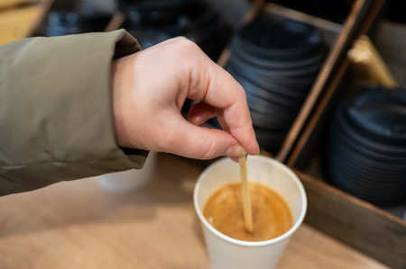 Close up of a female hand in green jacket mixing fresh cappuccino or latte with a disposable stirrer...